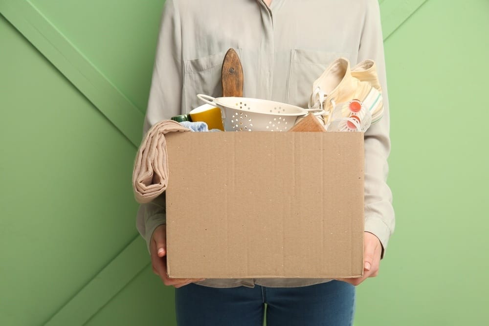 femme tenant une boîte remplie d'ustensiles de cuisine contre un mur vert